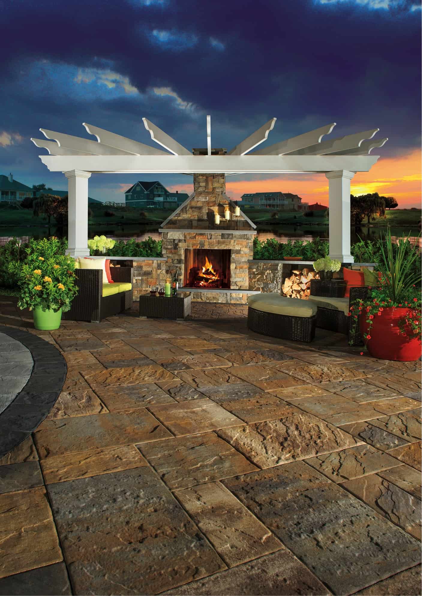 A cozy outdoor patio scene at sunset, featuring a stone fireplace with a lit fire under a white pergola. Surrounding the fireplace are wicker chairs with cushions, potted plants on meticulously laid paving stones from Nassau County, and a vibrant sky transitioning from a radiant sunset to a darkening evening.