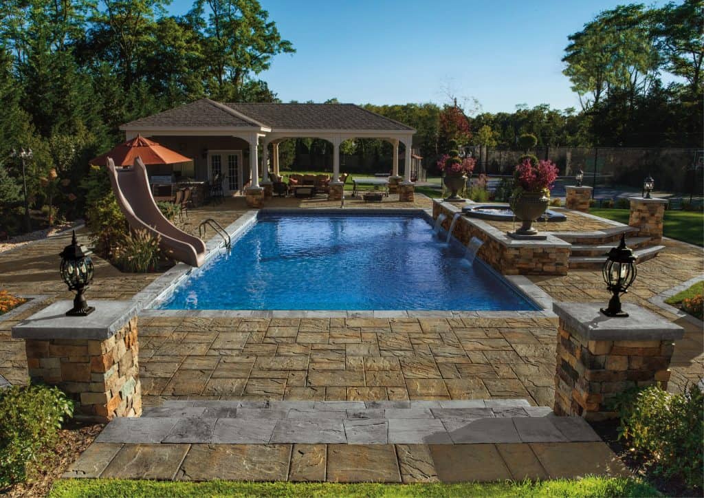 A backyard pool area with a rectangular swimming pool, stone steps and pillars, a waterslide, and a poolside pavilion with seating. Surrounding the pool are well-maintained paving stones from Nassau County and lush greenery. The setting is serene and inviting.