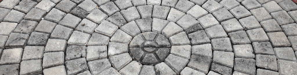 A close-up photo of a circular cobblestone pattern, featuring paving stones from Queens County. The stones are arranged in concentric rings, radiating outward from a central stone in the middle. They are primarily shades of gray, with a weathered and slightly rough texture.