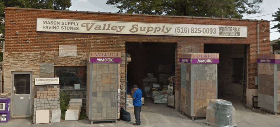 A man in a blue shirt stands outside Valley Supply, a masonry supply store in Nassau County with an open garage displaying stacks of paving stones and building materials. The building has a brick facade and signage with a contact number and details about their extensive masonry supplies.