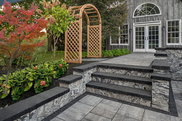 A well-landscaped backyard features stone steps leading to a raised patio area with decorative railing. A wooden trellis arch adorned with greenery stands along a pathway, and the house facade includes large glass doors and a semicircular window, showcasing the beauty of masonry supplies in Nassau County.