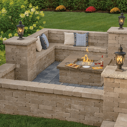 A cozy outdoor patio with stone walls and corner seating, adorned with cushions. A fire pit with dancing flames is in the center, surrounded by snacks and drinks. Decorative lanterns and blooming flowers enhance the tranquil garden ambiance, all crafted with quality masonry supplies from Queens County.
