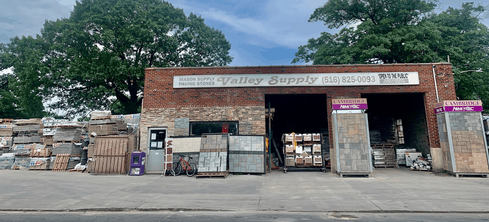 A brick building with the sign "Valley Supply" and contact information above the entrance. The lot is filled with stacks of various construction materials, including bricks, pavers, and masonry supplies for Nassau County. Two Cambridge Pavingstones displays are situated on either side of the entrance. Trees are visible in the background.