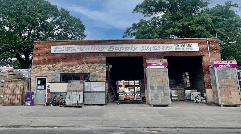 A masonry supply store called Valley Supply, with signs reading "Mason Supply," "Paving Stones Nassau County," and contact information. The entrance is flanked by stone displays and stacks of materials. A bicycle is parked on the left, and trees are visible in the background.