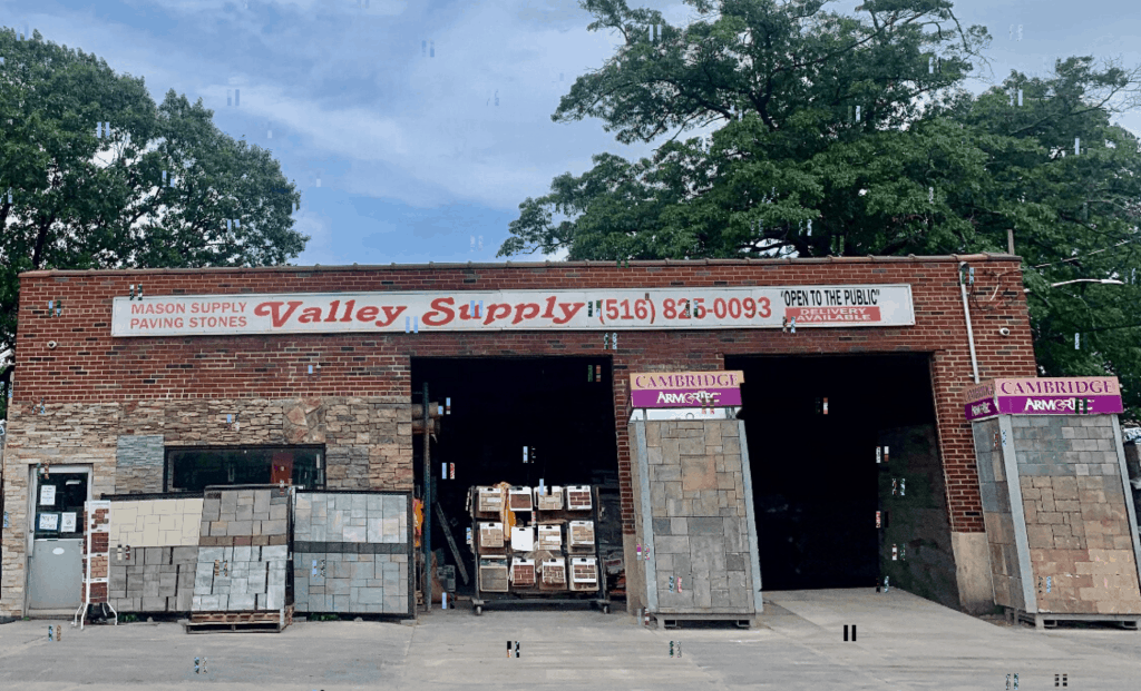 A brick building with a sign that reads "Valley Supply" and a phone number. The storefront, showcasing various paving stones Nassau County residents trust, reveals more stone products through two open garage doors. Trees are visible in the background under a clear sky.