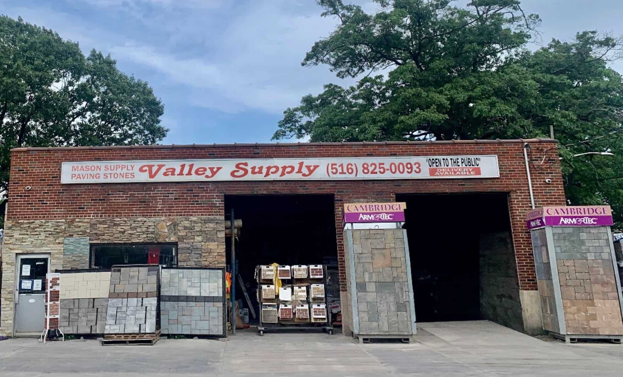 A brick and stone building with a sign that reads "Valley Supply," along with a contact number, stands prominently. Various paving stones and supplies are showcased both outside and inside the building. The entrance is flanked by racks of stones, with trees in the background—your go-to for masonry supplies in Queens County.