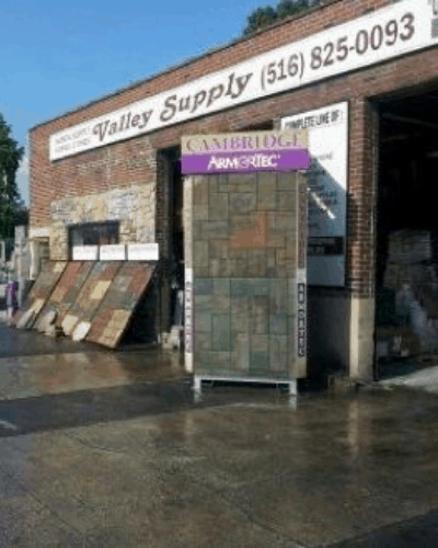 A brick building with a sign reading "Valley Supply" and a phone number above the entrance. Various stone and tile samples, including top-notch masonry supplies Nassau County, are displayed outside. A stand with samples labeled "Cambridge ArmorTec" is prominently placed near the entrance.