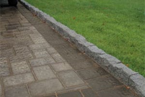 A wet stone-paved pathway, crafted using premium masonry supplies from Queens County, is bordered by a low, gray stone curb on the left. On the opposite side of the curb, there is a neatly trimmed green lawn. The pathway is partially visible, extending into the distance.