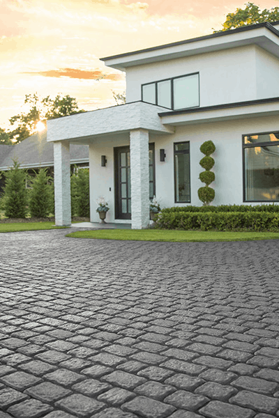 Modern white house with large windows at dusk, featuring a flat roof and clean lines. The entrance is framed by stone columns, flanked by decorative greenery. The foreground showcases a cobblestone driveway leading up to the house, built using quality masonry supplies from Nassau County.