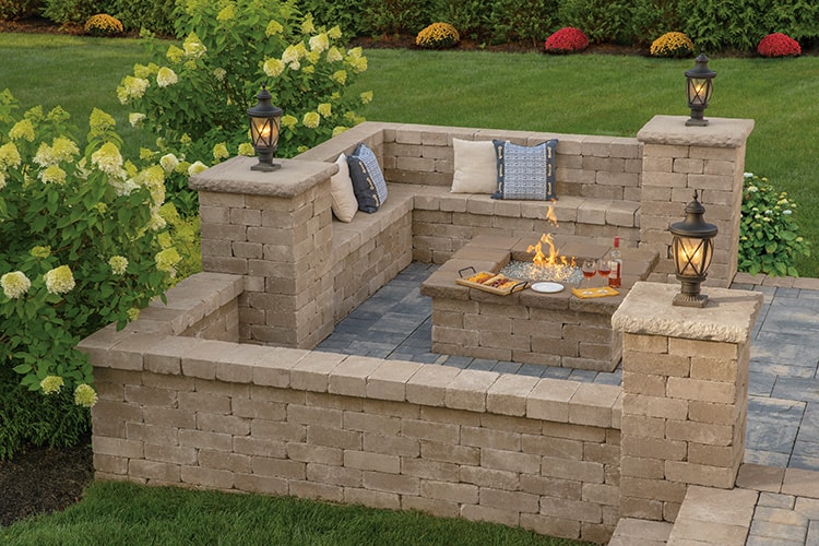 A cozy outdoor patio with stone walls and built-in bench seating, featuring cushions and pillows. A rectangular fire pit occupies the center, surrounded by snacks and drinks on a table. Decorative lanterns adorn the walls, with lush greenery and flowers in the background. The paving stones from Queens County add a charming touch.