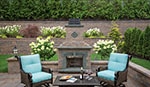 A cozy outdoor patio with two cushioned chairs facing a stone fireplace crafted from fine masonry supplies Nassau County offers. A small table between the chairs holds a bottle of wine and two glasses. The area is surrounded by lush greenery, blooming flowers, and a well-maintained lawn, providing a serene garden atmosphere.