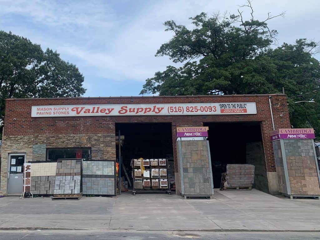 A building with a sign that reads "Valley Supply" and contact information. The entrance showcases various paving stones Nassau County and construction materials. Trees adorn the background, while the clear pavement in front highlights the masonry supplies Nassau County offers. A "Cambridge" sign is visible above the stone displays.