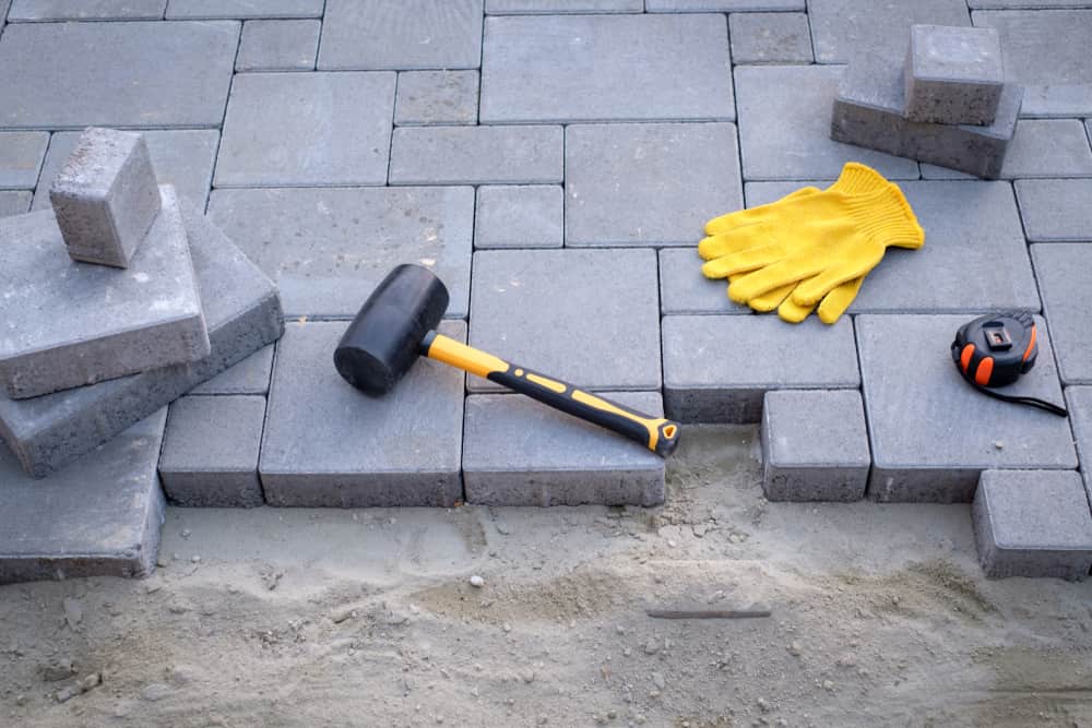A work in progress of a patio being paved with dark gray rectangular bricks. A rubber mallet, bright yellow work gloves, and a tape measure are placed on top of the bricks while some tools rest on the sand foundation. The finest masonry supplies in Queens County contribute to this meticulous project.