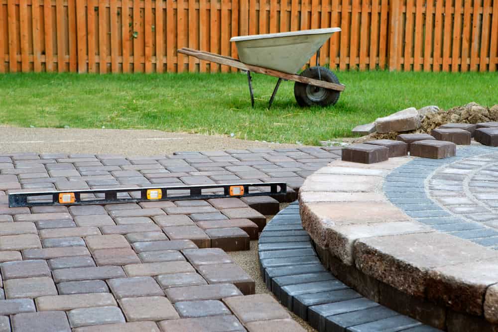 A level tool is placed on a partially completed paved surface in a backyard. There are various paving stones from Nassau County neatly arranged, and a wheelbarrow sits on the lawn next to a wooden fence in the background.