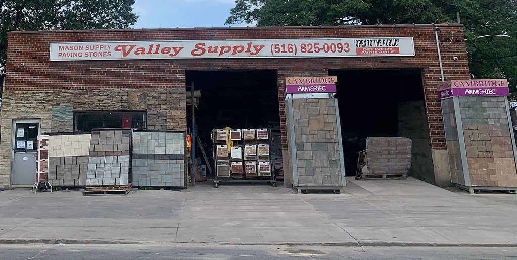 A brick building with a sign reading "Valley Supply" and "Mason Supply, Paving Stones" above the entrance. Open to the public, it showcases different types of paving stones and several pallets of materials inside the open garage. For top-quality masonry supplies in Nassau County, this is your go-to spot.