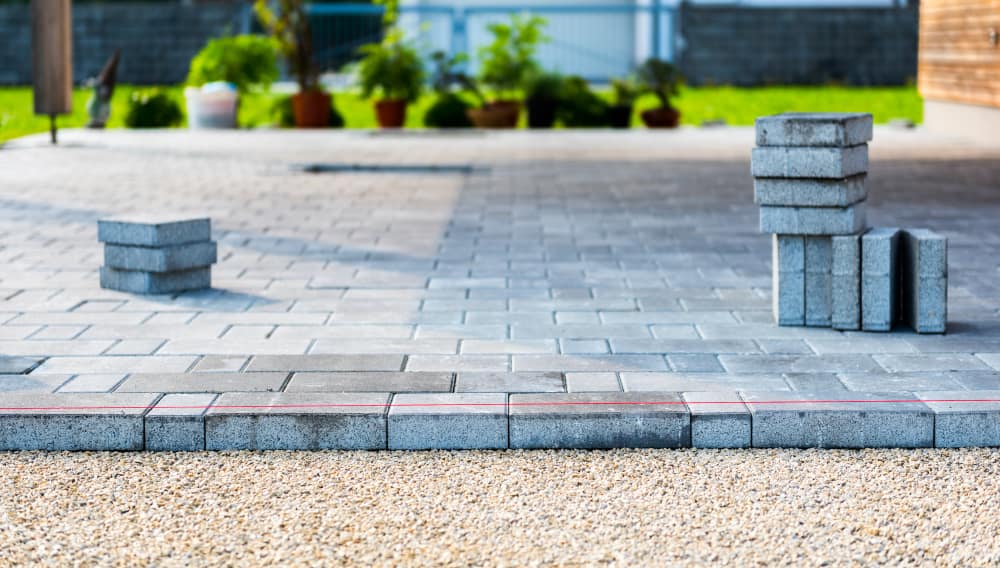 A paved patio is under construction with stacks of concrete blocks from Masonry Supplies Queens County positioned on it. The patio has an unfinished border with gravel and a red guideline. In the background, there are potted plants and a lush green lawn.
