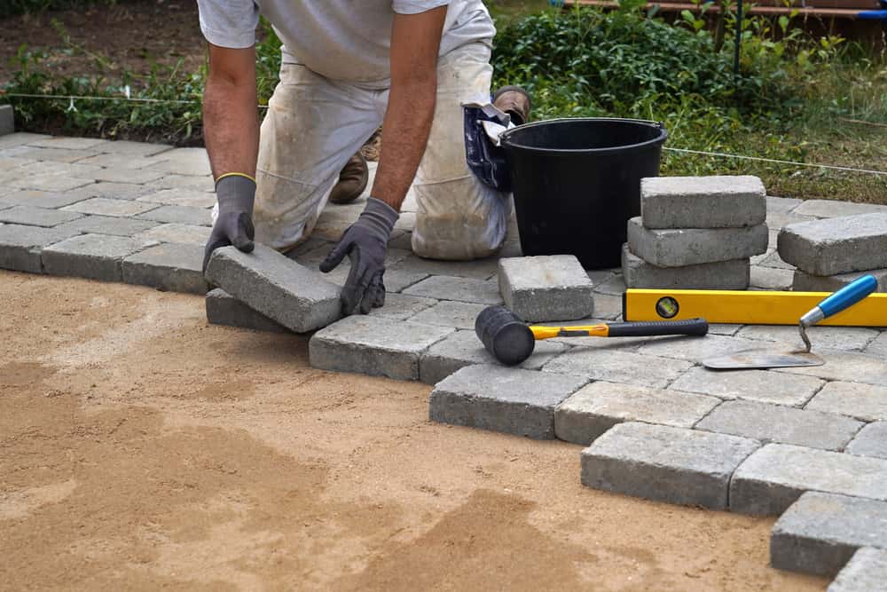 A craftsman lays concrete paving stone blocks on sand.