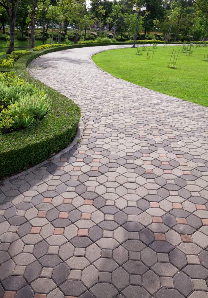 A winding park pathway paved with hexagonal stones, some accented in red. Lush green lawns and trimmed hedges line the path, with tall trees and street lamps visible in the background. The scene is tranquil, inviting for a peaceful walk, showcasing craftsmanship akin to Paving Stones Queens County.