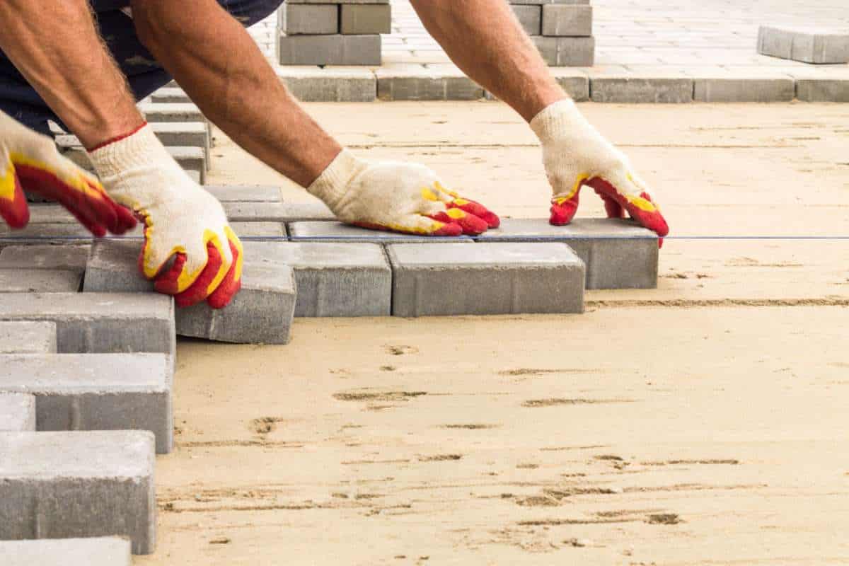 Workers wearing gloves carefully laying gray paving stones in a sand base, forming a pavement. Several other paving stones are stacked nearby, ready for installation. The high-quality Paving Stones Nassau County ensures durability and flawless finish for any outdoor project.