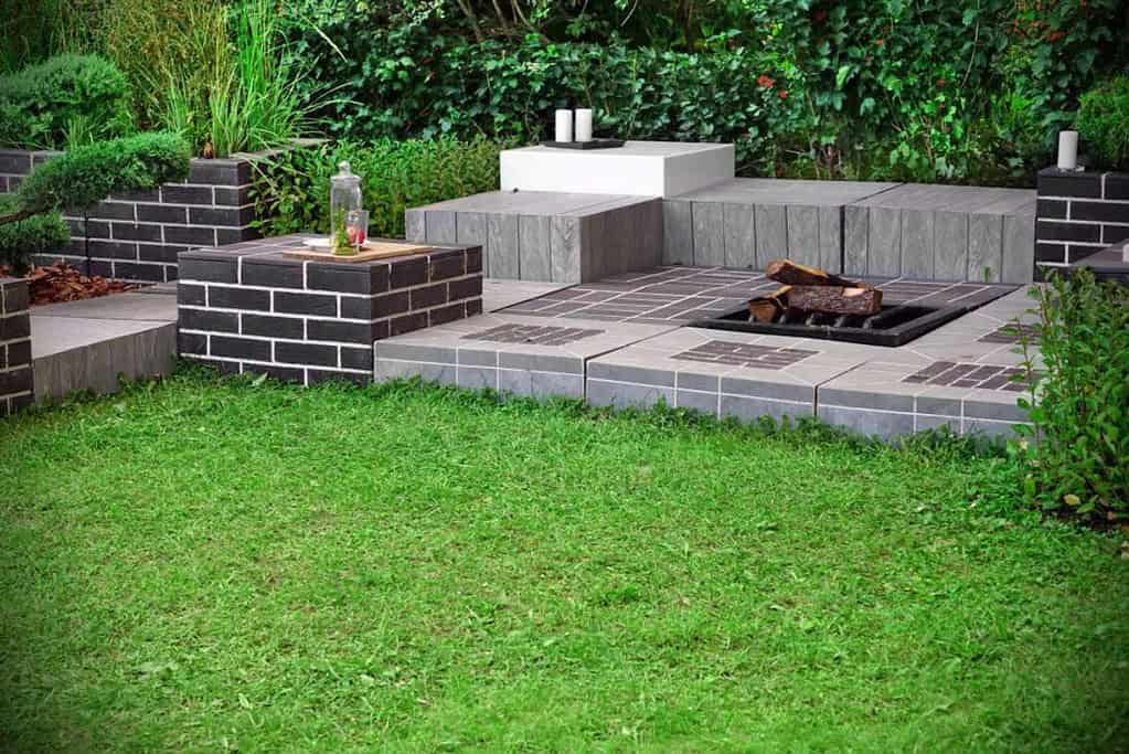A modern outdoor seating area with a fire pit features multiple levels of square and rectangular paving stones from Queens County. Green grass surrounds the space, and lush foliage can be seen in the background, creating a serene atmosphere.