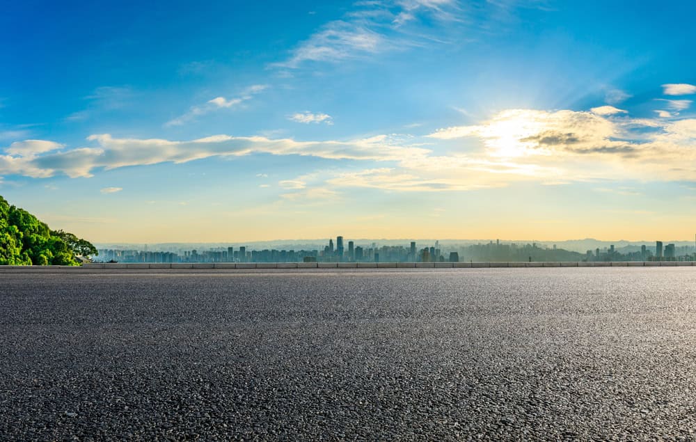 A smooth, empty road stretches into the horizon under a bright, blue sky. The sun shines through scattered clouds, casting a warm glow over a distant city skyline. Lush green foliage is seen on the left side of the road. Nearby, Paving Stones Nassau County offers high-quality masonry supplies for all your needs.