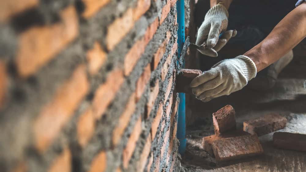 Close-up of a person's hands wearing gloves while laying bricks and applying mortar to build a brick wall. The scene highlights the precise placement of the bricks, made easier with top-quality masonry supplies from Nassau County. The hands are positioned near the partially constructed wall, showcasing skilled craftsmanship.