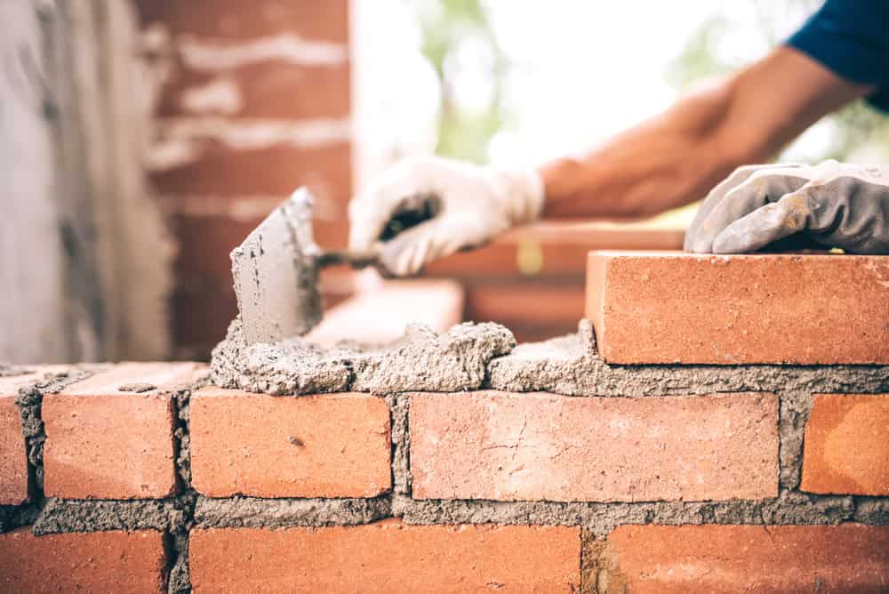 A pair of hands wearing white gloves is seen constructing a brick wall. One hand holds a trowel and spreads mortar onto the orange bricks, while the other hand steadies them. Using top-quality masonry supplies from Nassau County, the wall is partially built, and another brick layer is being added.