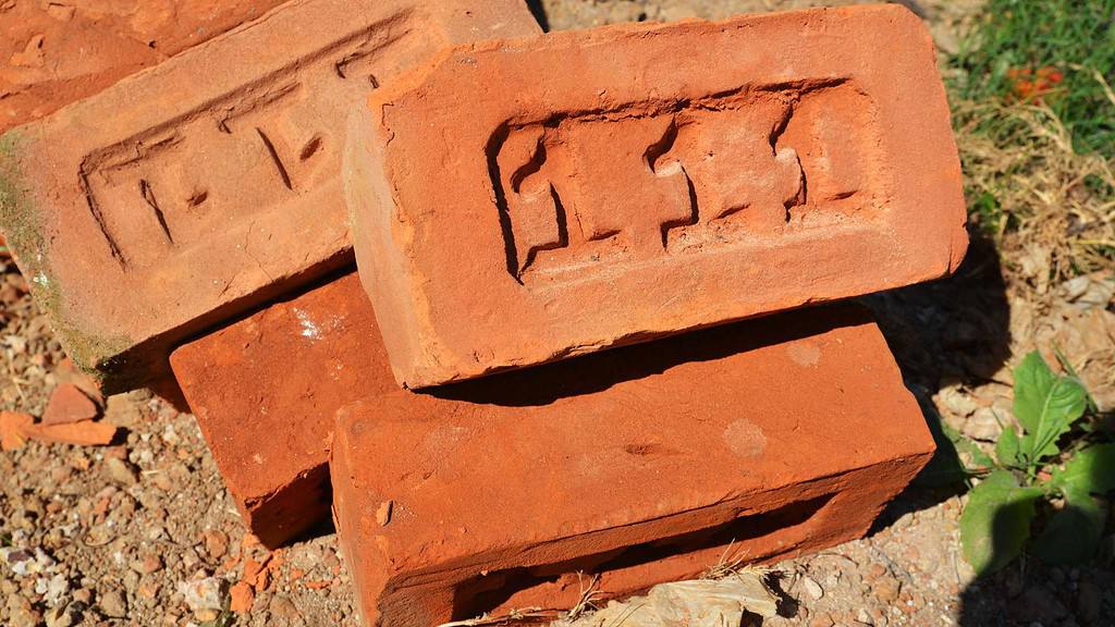Three red clay bricks stacked on the ground. Two bricks have rectangular indents with indistinct markings inside them. The bricks are surrounded by dirt and small pieces of debris. A small green plant is partially visible in the corner, reminiscent of masonry supplies in Nassau County.