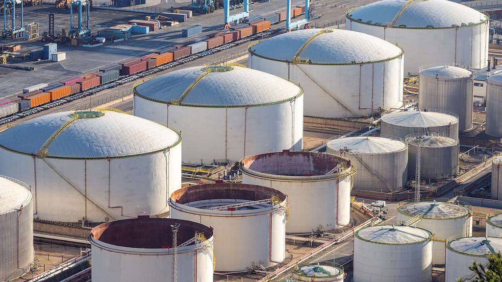 Aerial view of numerous large, white, cylindrical storage tanks with domed roofs at an industrial site. Surrounding infrastructure includes pipelines and rectangular containers, resembling those found in a refinery or a facility offering masonry supplies in Nassau County.