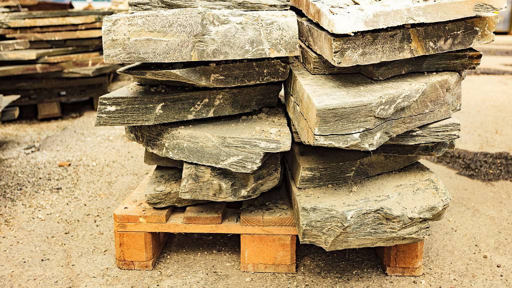 A wooden pallet holds a stack of uneven, large, rough-cut stone slabs of varying thicknesses. The slabs, perfect as paving stones in Nassau County, are weathered and dusty with irregular edges. The pallet is situated on a gravelly surface in an outdoor setting.