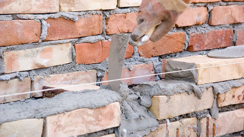 A close-up of a bricklayer using a trowel to apply mortar between bricks from masonry supplies in Nassau County while constructing a brick wall. A white string line is positioned horizontally across the bricks to ensure they are laid straight. The bricks vary in color from red to beige.