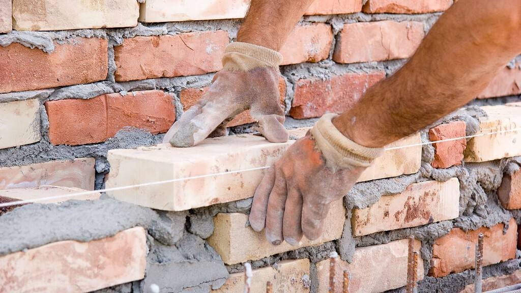 A close-up of two hands wearing gloves, positioning a new brick in place on a partially constructed brick wall. The mortar is visible between the bricks and a string line is used for alignment, showcasing the quality masonry supplies Queens County offers.