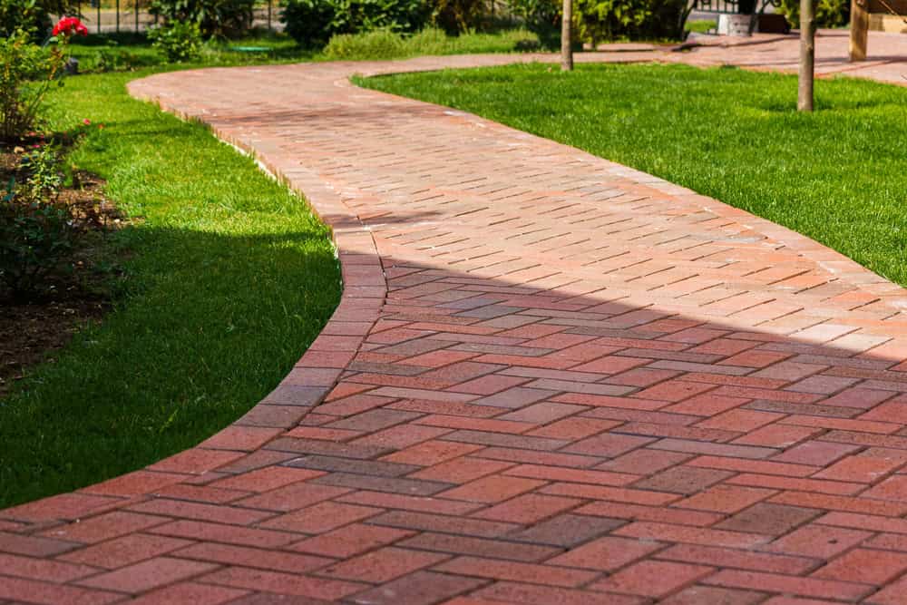 A winding brick pathway curves through a well-maintained green lawn, much like those created with quality paving stones found in Nassau County. The red and brown herringbone-patterned bricks contrast with the lush grass. The surroundings include some plants and bushes, adding to the serene outdoor setting.