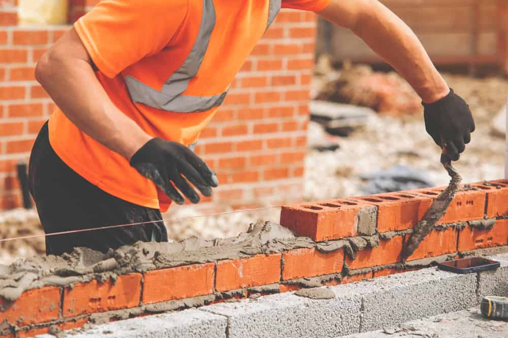 A construction worker wearing an orange shirt and black gloves is laying bricks with mortar to build a wall. A string line is visible, ensuring alignment, and the wall is partially constructed with red bricks. The background shows more construction materials from Masonry Supplies Nassau County.